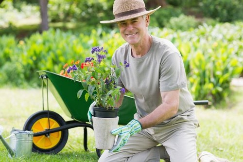A middle-aged man dressed in casual gardening attire, including a wide-brimmed straw hat, light grey t-shirt, and gardening gloves, is kneeling on a well-maintained lawn in a garden setting. He is holding a potted flowering plant with purple and red blooms, which appears to be freshly picked or prepared for planting. Behind him, a green wheelbarrow with bright yellow wheels is positioned on the grass, partially filled with gardening tools and supplies, indicating outdoor maintenance activities. The background features a lush, green hedge and trees, creating a natural boundary for the garden space, with vibrant greenery and a slightly blurred garden environment visible in the distance. The scene is illuminated by natural daylight, suggesting a clear, sunny day, suitable for gardening work. This setting reflects typical garden care and maintenance, emphasizing professional landscaping and outdoor plant care services offered by Hedge Trimming Brompton in the Brompton area, contributing to well-kept outdoor spaces in the locality.