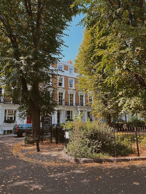 An exterior view of a garden area in front of a multi-story white Victorian-style building on Exhibition Road, Brompton. The garden features a variety of lush green trees with dense foliage, casting d
