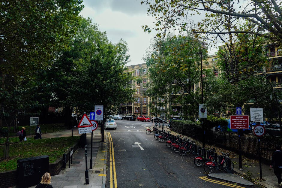 An exterior view of a garden area in front of a multi-story white Victorian-style building on Exhibition Road, Brompton. The garden features a variety of lush green trees with dense foliage, casting d