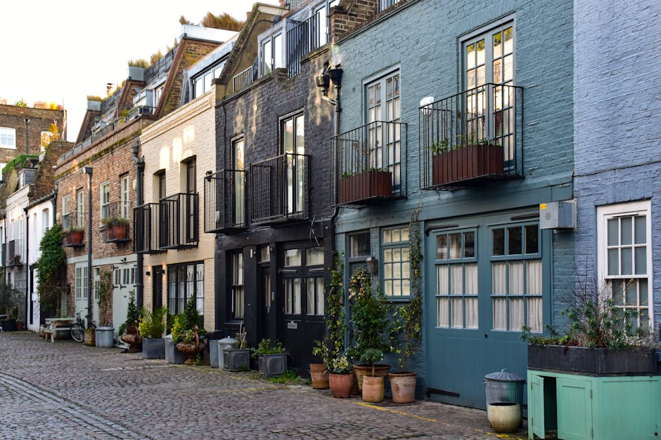 A row of charming, narrow terraced houses along Brompton Road mews, each featuring a small balcony with black metal railings and potted plants. The houses are painted in various pastel and neutral sha