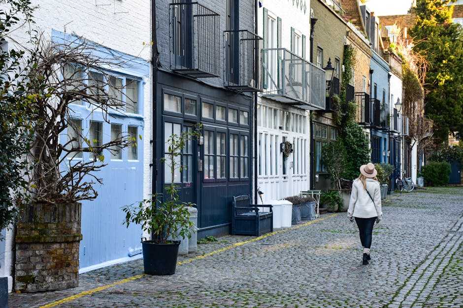 A row of charming, narrow terraced houses along Brompton Road mews, each featuring a small balcony with black metal railings and potted plants. The houses are painted in various pastel and neutral sha